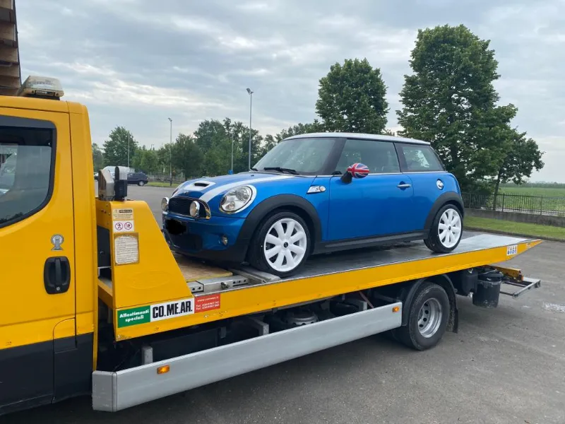 Carro attrezzi giallo con pianale che trasporta una utilitaria blu con tetto bianco